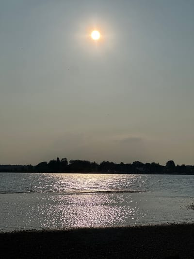 The photograph captures a serene lake scene with the sun shining brightly in a hazy sky. The sun's rays create a shimmering, sparkly effect on the water's surface. In the background, a silhouetted line of trees and structures forms a distant shoreline. The foreground is a dark, pebbly beach. This image was taken in Chieming, Germany, likely near the shore of Chiemsee, a large lake in Bavaria. The overall impression is one of a peaceful, late afternoon or early evening setting.