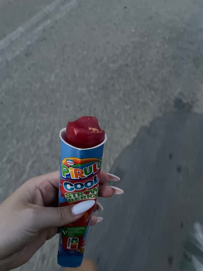 A hand with long, white-polished fingernails holds a Pirulo Cool Strawberry ice pop. The ice pop wrapper is blue and red with colorful text that reads "PIRULO COOL STRAWBERRY". The ice pop itself is bright red and appears to be partially melted. The background is a gray, textured surface, likely pavement. The scene is outdoors, possibly in the city of Skala Avdiron, Greece, indicated by the context.