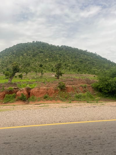 The image depicts a natural landscape with a focus on a verdant hillside leading up to a mountain. In the foreground, there is a paved road with a single yellow line dividing the lanes, bordered by a gravel shoulder. The hillside is characterized by reddish-brown earth in some areas, with sparse vegetation and a few scattered trees. Further up the slope, the terrain becomes more densely covered in green foliage and trees, extending to the summit. The sky is overcast with a thick layer of clouds. The scene suggests a rural or semi-rural environment, potentially on the outskirts of a settlement. The location is identified as Minda, Nigeria. There are no people or identifiable man-made structures other than the road. The overall impression is one of a natural, undeveloped landscape.