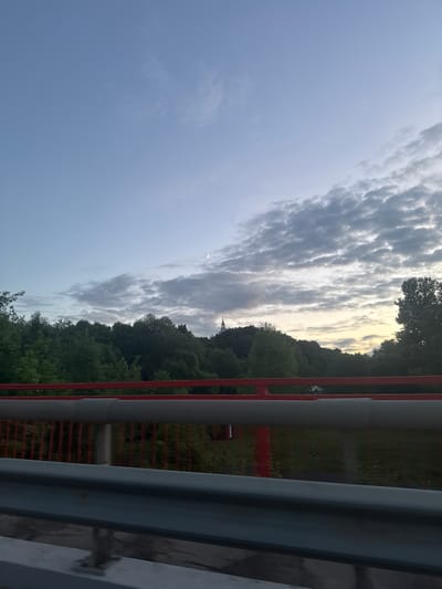 The image shows a view from a bridge in Vereya, Russia, overlooking a tree-covered hillside. The sky is a pale blue with scattered clouds, and a sliver of the moon is visible. A tall, slender spire, likely belonging to a church, rises from the treeline in the distance. The foreground of the image is dominated by the red railing and grey metal barrier of the bridge. The scene appears to be captured during twilight or early evening. There are no people or animals visible in the image.