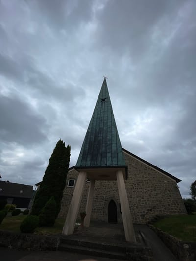 This image depicts a church in Schorbach, Germany. The church features a stone facade and a tall, pointed spire covered in a weathered, greenish-blue material. The spire is supported by angled concrete pillars that extend from the ground to the base of the spire's overhang. A dark, arched doorway is visible on the stone wall beneath the spire. Adjacent to the church, on the left, is a tall, slender evergreen tree. In the background, residential buildings with tiled roofs can be seen. The sky is overcast with heavy, grey clouds. The foreground shows a paved walkway leading to the church entrance, flanked by steps and stone walls. The overall scene suggests a cloudy or impending stormy day.