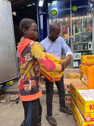 The scene depicts two males engaged in handling merchandise in a street-level commercial area in Ajah, Nigeria.
The individual on the left is a young male, appearing adolescent, with dark skin and short, dark, tightly curled hair. He wears a multi-colored hooded sweatshirt featuring a blend of red, yellow, black, and orange, patterned with various graphics including "GUCCI," "VERSACE" medusas, and two stylized depictions of the artist Drake. He is also wearing dark pants with blue vertical stripes. His gaze is directed to the right. He is holding a large, yellow cardboard box.
The individual on the right is an adult male, with dark skin and short, dark hair. He wears a light blue button-up shirt with sleeves rolled up, dark trousers, and dark sandals. He is positioned facing the yellow box and is also holding it, suggesting a joint effort in moving or transferring the item. His gaze is directed downwards towards the box.
The yellow cardboard box being handled is labeled "KING'S VEGETABLE OIL," "COOKING and SALADS," and indicates a quantity of "6x2L." Multiple identical yellow boxes are stacked on the paved ground to the right of the individuals, alongside some brown cardboard boxes and large orange plastic containers.
In the background, a blue-framed glass storefront is visible, displaying various packaged items, possibly electronics or small appliances. Two "oraimo" logos are affixed to the glass. A white rectangular light fixture is mounted above the storefront entrance. To the far left, a partially open grey metal door or security shutter is visible. The ground is a paved surface, exhibiting minor debris.