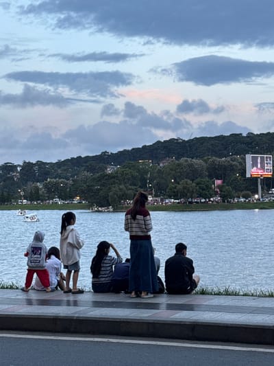 The image depicts a scene at Xuan Huong Lake in Da Lat, Vietnam. In the foreground, a group of people, including children and adults, are sitting and standing by the edge of the lake. The lake's surface reflects the overcast sky and the green, tree-covered hills that form the backdrop of the city. On the distant hillside, buildings and trees are visible, with a large billboard displaying a live broadcast on the right side. Several swan-shaped paddle boats are scattered across the water. The sky is filled with a mix of gray and light blue clouds, with hints of pink suggesting either dawn or dusk. The overall atmosphere is calm and serene, with people enjoying the view of the lake and the surrounding scenery.