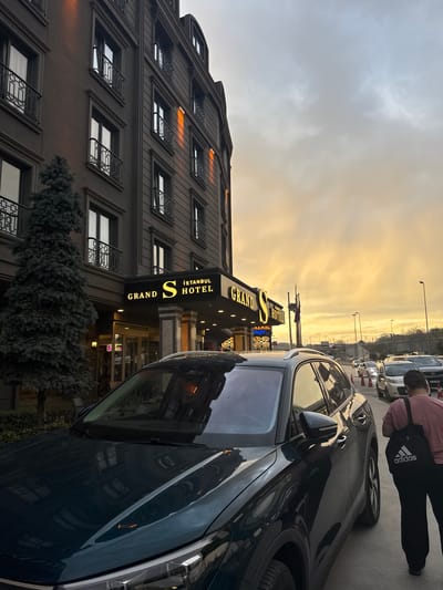 A dark-colored multi-story building, identified as the "Grand S Istanbul Hotel," stands prominently in Esenler, Turkey, under a sky illuminated by the golden-orange light of sunset or sunrise. The hotel's facade is dark gray or brown, featuring multiple windows with black decorative balustrade balconies. The hotel's illuminated gold sign, "GRAND S ISTANBUL HOTEL," is positioned above the entrance. To the left of the entrance, a tall, dark green coniferous tree stands on the sidewalk.

In the immediate foreground, a dark green SUV is parked, with its hood and windshield reflecting the building and sky. A person, viewed from behind, walks on the sidewalk next to the SUV. This individual wears a reddish-brown t-shirt and dark trousers, carrying a black Adidas drawstring bag over their left shoulder.

The street extends to the right, showing additional parked vehicles and a line of orange traffic cones. In the background, an elevated road or bridge structure is visible, supported by pillars, with streetlights lining its edge. The overall illumination is a mix of the natural golden sky light and artificial lights from the hotel and streetlights.