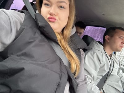 The image depicts three individuals inside a vehicle. Two are visible in the front seats: a woman with blond hair and a black puffer jacket, and a man wearing a light grey jacket. Both are wearing seatbelts. A third person is partially visible in the reflection in the front window of the vehicle, behind the front passenger seat. The scene takes place inside a car, presumably in Ledukhi, Russia, as this information was provided. The weather seems to be cold or overcast, suggested by the jackets worn by the occupants. The interior of the vehicle is primarily grey and black. Outside the window, bare trees are visible, suggesting it is either late fall, winter or early spring. The individuals are seemingly traveling in the vehicle.