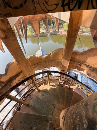 An overhead shot reveals a weathered spiral staircase in the city of Thủy Xuân Ward, Vietnam. The staircase's center column is heavily marked with graffiti. The treads of the staircase are textured. A rusty metal railing runs along the inner edge of the staircase. Outside the staircase, a moat-like body of water is visible, bordered by architectural features resembling large teeth or spikes. Graffiti is also visible on these exterior walls. The lighting is bright, casting stark shadows across the scene. The overall impression is one of decay and neglect within an unusual architectural setting.