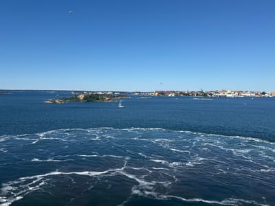 The image shows a wide view of a body of water, likely a harbor or archipelago, with a city skyline in the distance. The foreground is dominated by the turbulent wake of a boat, characterized by frothy white lines and churned water, indicating movement. Beyond this, the water is a deep blue, with smaller ripples. In the middle ground, several small islands are visible, some with buildings and vegetation. A white sailboat is present on the water. The background features a city with a varied architecture, including red-roofed buildings, taller structures, and cranes, suggesting an urban port area. The sky is a clear, bright blue with a single white bird flying high. The scene appears to be Karlskrona, Sweden, given the characteristic archipelago environment and urban port structures.