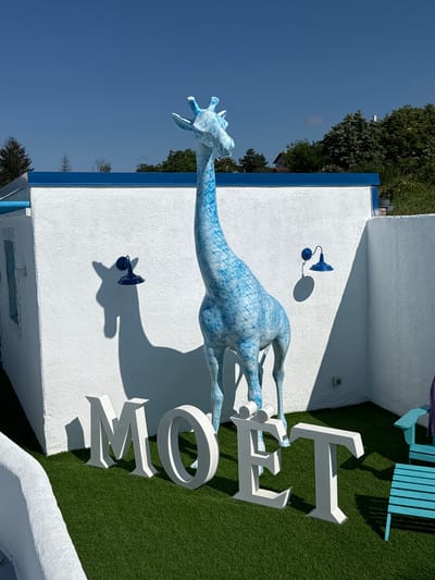 The image depicts an outdoor scene in Baliovtsi, Bulgaria, under a clear blue sky. A large, blue and white speckled giraffe statue stands prominently in the center of the frame. In front of the giraffe, large white letters spell out "MOET" on a patch of artificial grass. To the right of the statue, a blue adirondack chair with a purple towel draped over it is visible. Two dark blue wall-mounted lamps are fixed to a white textured wall behind the giraffe, one casting a distinct shadow of the giraffe's head and neck. The environment appears to be a well-maintained outdoor area, possibly a garden or patio, with white stucco walls surrounding the space.