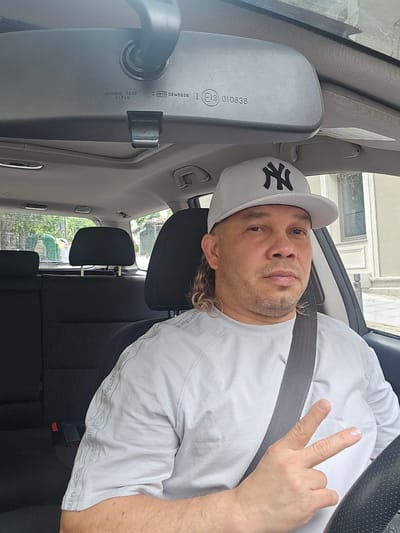 An adult male is prominently featured in a portrait-style shot taken from inside a car, likely from the passenger's perspective. The image, originally rotated, shows the man seated in the driver's seat, secured by a dark grey seatbelt across his chest and lap. He has medium-length curly or wavy brown hair visible beneath a light grey baseball cap adorned with a black "NY" logo, characteristic of the New York Yankees. His attire includes a light grey short-sleeved t-shirt, which features a repeated "Umbro" brand logo along a band on the shoulder/collar area. He looks directly towards the camera with a neutral to slightly serious expression. His left hand is raised, making a peace sign with his index and middle fingers.
The car's interior is visible, including parts of a black textured steering wheel, black fabric seats, and headrests. A prominent rearview mirror hangs in the upper part of the frame, displaying manufacturing text such as "ICHIKOH 8288", "JAPAN", and a certification mark "E13 02*0836". Through the car windows, an outdoor scene is discernible, featuring bright natural light, glimpses of buildings with windows, and some green foliage, suggesting an urban or suburban setting. The bright lighting indicates it is daytime. The scene is located in Real Sitio de San Ildefonso, Spain.