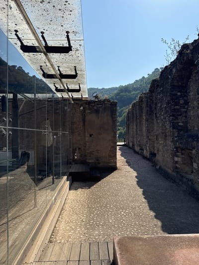 The image shows a view of the Doria Castle in Dolceacqua, Italy. In the foreground, a modern glass structure with a speckled roof is reflected in its glassy facade. To the right, the ancient stone ruins of the castle walls form a pathway that recedes into the distance. The pathway is paved with small stones and is partially covered in shadow. In the background, a lush green hillside dotted with trees rises towards a clear blue sky. The scene is bathed in bright sunlight, casting sharp shadows.