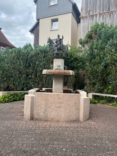 The image shows a bronze sculpture of two figures on horseback, mounted on a granite fountain. The sculpture depicts one figure appearing to lead, with the other figure following closely behind. The fountain itself is constructed of speckled pink and grey granite, with carved basins. Inscribed on the granite base of the fountain is text, which appears to read "A.D. 2004 500 JAHRE ST. TRINITAIS". The fountain is situated outdoors, with a backdrop of lush green trees and hedges. To the left of the fountain, a building with a grey, tiled roof is visible, suggesting an urban or suburban setting. The sky is overcast, indicating a cloudy day. The scene is calm and appears to be a public space, possibly a park or a square in Neustadt, Germany.
