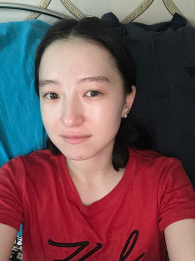 A young East Asian female is lying down, facing the camera. She has dark hair pulled back from her face and is wearing a red t-shirt. She has a small pearl earring in her left ear. Behind her, a textured blue fabric is visible on the left, and a dark fabric is visible on the right. The background appears to be a wall or headboard with a decorative pattern. The image is a selfie, taken from a slightly overhead angle. The overall lighting is dim, with some areas brighter than others. The location in Nookat, Kyrgyzstan, cannot be determined from the image alone.
