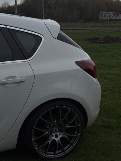 The image shows the rear quarter of a white car parked on a grassy area. The car features a dark-colored wheel with a multi-spoke design. A red tail light is visible. The background consists of grass and what appears to be trees and a building in the distance. Without further data, it is not possible to confirm that the picture was taken in Semeno-Aleksandrovka, Russia.