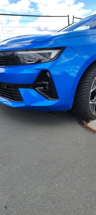 The image shows the front driver's side of a bright blue car parked on asphalt, likely in Hattersheim, Germany. The sky with scattered clouds is reflected in the car's paint. The car's headlight, fog light, and grill are visible. A tire and a curb edge are positioned at the upper right. A metal fence is at the left of the car.