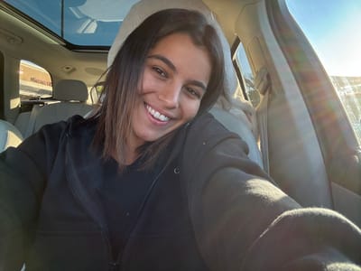 A young adult female is depicted from the chest up, positioned inside the passenger compartment of a vehicle. She has shoulder-length dark brown hair, is smiling broadly at the camera, and is wearing a black ribbed top under a black zippered hoodie. The vehicle features light-colored leather seating and a panoramic sunroof, reflecting a clear blue sky. Bright sunlight enters the vehicle from the right, producing lens flare. Through the rear left window, a tan commercial building with a partial "Targ..." sign is visible, identifying it as a Target store. The scene is located in a parking lot adjacent to the Target store at 2700 Brunswick Pike, Lawrence Township, New Jersey, United States, under natural daylight conditions.