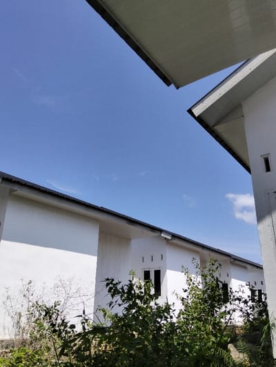 A bright sky fills most of the frame, peeking between roof overhangs of white buildings. One building is close, partially obscuring the sky to the right. The other buildings are a line of white, low structures with dark gray rooflines. Some windows are visible. Weeds and foliage fill the foreground, blocking the lower part of the buildings.