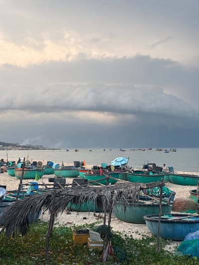 The image depicts a beach scene in Thuận Thành, Vietnam, with numerous traditional Vietnamese basket boats and fishing boats scattered across the sandy shore and in the water. The sky is overcast with dramatic, dark clouds, suggesting approaching rain or stormy weather.
In the foreground, several green and blue basket boats are pulled up onto the sand. Some of these boats are empty, while others contain fishing nets, equipment, and what appear to be bundles of supplies. A makeshift shelter made of palm fronds covers some of the boats, offering shade. Green vegetation grows along the edge of the beach in the lower part of the image.
Further out on the water, a larger number of fishing boats are visible, some appearing to be actively engaged in fishing activities. The coastline stretches into the distance on the left side of the image, with buildings and a rocky outcrop visible.
Two individuals are present on the beach. One person is seated in a green basket boat near the center of the image, while another person is standing near a boat on the right side. The overall impression is a working fishing village preparing for or experiencing inclement weather.