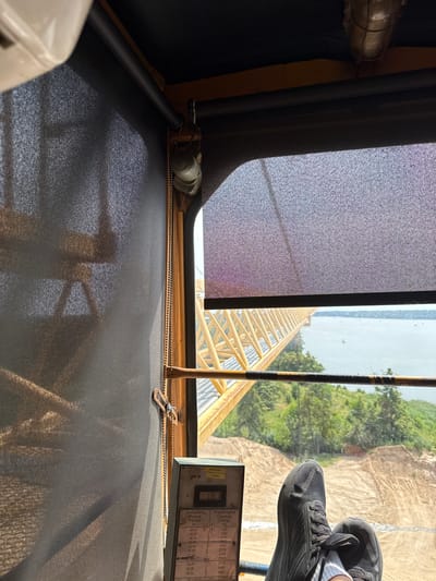The image is taken from the perspective of someone sitting in the cab of a crane, looking out a window. In the foreground, a person's black athletic shoes are resting on the window ledge or a similar surface. The shoes appear to be worn. Through the window, a yellow crane boom is visible extending over a body of water, likely Lake Mikołajskie, given the context of Mikołajki, Poland. Trees and sandy terrain are visible on the opposite shore. A control panel with some gauges and text in what appears to be Polish and German is also visible on the right side of the frame. The overall scene suggests a construction or industrial site near a body of water.
