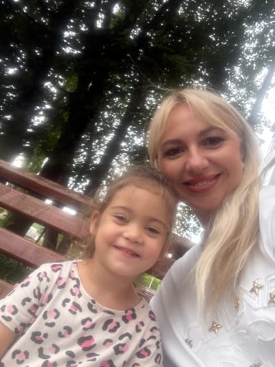 In Ishimbay, Russia, a woman with blonde hair and a young child with blonde hair are seated on a wooden bench. The woman is wearing a white top with star embellishments, and the child is wearing a white t-shirt with a pink and black leopard print. Both are smiling and looking at the camera. They are outdoors, with trees and dappled sunlight in the background. The bench appears to be made of dark reddish-brown wood with visible grain. The overall scene suggests a casual outdoor setting.