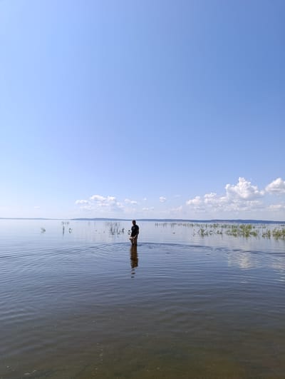 A person stands in shallow water on a bright, clear day. The person is wearing a dark shirt and light-colored shorts, and their legs are submerged to the knee. The water is calm, with ripples spreading out from the person's position, reflecting the blue sky. In the background, a distant shoreline is visible, lined with vegetation. The sky is a vast expanse of clear blue, with a few scattered white clouds near the horizon. The overall scene conveys a sense of peacefulness and solitude in a natural environment. The location is the city of Vekoshinka, Russia.