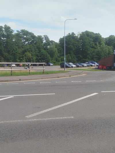 The image captures a scene in Cwmbran, United Kingdom, focusing on a section of a road and a parking lot adjacent to a wooded area. In the foreground, a grey asphalt road is visible with white lane markings, including a solid white line and broken white lines, indicating traffic flow. A section of a sidewalk with yellow lines runs along the edge of the road.
Behind the sidewalk, a wooden fence with multiple horizontal rails separates the road from a parking lot. The parking lot contains several cars parked in a row, including a blue car, a silver car, an orange car, a white car, a black car, and a dark grey car. The cars are positioned on a grey paved surface.
Further back, a dense line of green trees forms a natural backdrop to the parking lot. A tall, grey lamppost with a light fixture stands near the parking lot, extending upwards towards a bright, cloudy sky. To the right of the parking lot, a brick building is partially visible, with red and black bins placed in front of it. The overall environment suggests a public area, possibly near a commercial or residential zone. There are no people visible in the image.