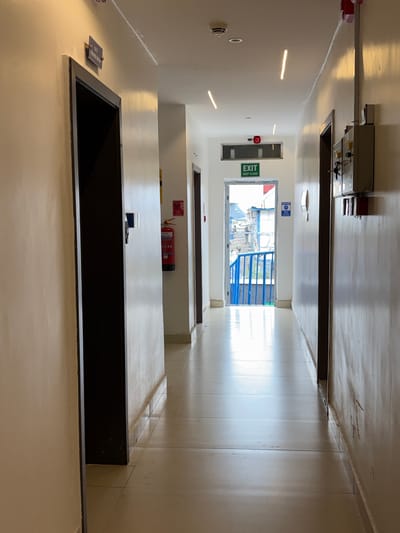 The image shows a long, dimly lit hallway with doors on either side. The hallway appears to be indoors, with fluorescent lighting along the ceiling. The walls are painted a light color, and the floor is tiled. At the end of the hallway, there is an open doorway leading to what appears to be an outdoor area with a blue metal railing. A green "EXIT" sign with white lettering is visible on the wall to the left of the open doorway. A red fire extinguisher is mounted on the wall in the middle ground to the right. There is a small rectangular sign with blue text on the wall to the left of the open doorway. The scene is static, with no people or activity visible. The overall impression is one of an ordinary building hallway.
The location context provided is Gwaska, Nigeria.