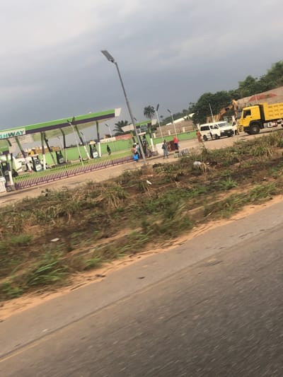 A roadside view shows a gas station with green and white signage, light poles, and palm trees. Several fuel pumps are visible. Beyond the gas station, there is a construction site with a yellow dump truck and a white crane truck. The foreground features a paved road and a grassy shoulder. The sky is overcast.