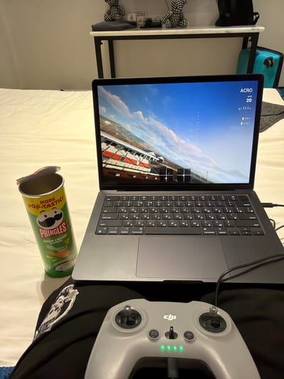 The image depicts a close-up of a person's lap with a drone controller in the foreground and a laptop open on a bed behind it. A partially opened can of Pringles chips is positioned to the left of the laptop. The laptop screen displays a first-person view of a drone flight, showing a racing track with red and white checkered barriers and a blue sky with clouds. The drone's altitude is displayed as 11.1 meters, and its speed is 20 km/h. The drone controller is grey and has various buttons and joysticks. Green LED lights indicate battery level. In the background, a white duvet covers the bed, and a shelf with decorative items and a turquoise suitcase is visible. The scene is indoors, suggesting a casual setting, possibly a bedroom or living area. The image does not provide any visual cues to indicate the location of Ban Phonsavang Neua, Laos.