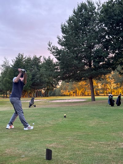A dynamic photograph captures a male golfer mid-swing on a golf course, likely in South Cave, United Kingdom, during the late afternoon or early evening golden hour. The golfer, positioned towards the right of the frame, wears a dark blue polo shirt, grey trousers, and white athletic shoes with dark stripes. He is shown with a golf club raised high, indicating either the top of his backswing or the start of his downswing. A white golf ball rests on a tee near his feet, with a black practice mat situated further right on the green grass.
The expansive golf course stretches behind the player, featuring a manicured green fairway and a distinctive sand bunker in the mid-ground. A golf push cart is visible behind the golfer, while two black golf bags filled with clubs stand in the far background on the right. Dense, tall trees with dark green foliage frame the left side of the image and line the course in the distance, where warm, golden sunlight dramatically illuminates the leaves. The sky above is a soft, pale blue transitioning to hints of purple, characteristic of sunset. The scene conveys a moment of focused athletic activity within a serene, beautifully lit natural setting.