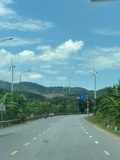 This is a landscape photograph taken from a moving vehicle on a paved road in Tân Lập Commune, Vietnam. The scene depicts a rural setting with lush green vegetation lining the right side of the road. In the background, rolling hills covered in trees rise up, and several wind turbines are visible on the slopes, indicating a wind farm. The sky is bright blue with scattered white clouds, suggesting it is daytime with pleasant weather.
The road itself is gray asphalt with white and yellow dashed lines marking the lanes. There are two motorcyclists riding on the road, heading away from the viewer. Along the side of the road, there are a few streetlights and a metal guardrail. Several road signs are visible, including yellow triangular warning signs and a blue rectangular sign with Vietnamese text. One of the visible signs reads "CHÚ Ý KHU DÂN CƯ" which translates to "Attention Residential Area" and below it "200m". Another sign appears to be a speed limit sign.
The overall impression is one of a scenic drive through a countryside area that utilizes renewable energy. The presence of the road, vehicles, and signs suggests this is an active route for transportation. The vegetation, including what appears to be banana trees, reinforces the tropical or subtropical location.