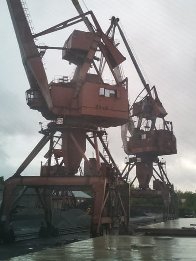 This image captures an industrial scene dominated by two large, aging gantry cranes, likely used for bulk material handling at a port or railway terminal. The cranes are painted a faded, reddish-brown and show signs of rust, indicative of their age and constant exposure to the elements.

The crane on the left features an operator's cabin with large windows and a sign bearing the number "52" in white with a red border. Its massive boom extends upwards and to the left. The crane on the right is similar in structure, also featuring an operator's cabin and a visible sign with the number "43". Part of a word, possibly "AN", is also faintly visible on the side of the right crane's cabin.

In the foreground, a calm body of water, possibly a canal or harbor basin, reflects the overcast sky and the lower parts of the cranes. The water's surface appears wet and still, suggesting either recent rain or a naturally calm condition. Beneath the cranes, extensive piles of dark, granular material, likely coal or aggregate, are visible. Railroad tracks run alongside these piles, indicating a system for loading or unloading materials from trains.

In the background, behind the industrial setup, some buildings are partially visible through scattered trees. The sky is overcast and bright, suggesting daytime with cloudy weather, contributing to a muted, industrial ambiance. No active loading or unloading operations are depicted; the scene appears momentarily static, showcasing the infrastructure itself. The overall setting strongly suggests a working port or a heavy industrial facility specializing in bulk cargo movement in Medvezhyegorsk, Russia.