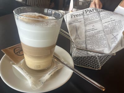 The image displays a close-up scene on a dark tabletop, likely within a café in Ribeauvillé, France. A tall, transparent glass occupies the left side, containing a layered coffee beverage. The drink exhibits four distinct layers: a light brown liquid at the bottom, followed by a medium brown layer, a thick white layer of steamed milk, and a substantial foamy white layer on top with a slight brown swirl. The glass is centered on a white, round ceramic saucer. A silver spoon rests diagonally on the saucer and extends onto the table. Two white packets of "SUCRE DE CANNE BIO" (Organic Cane Sugar) and one brown packet labeled "MANDALOU de provence" are also positioned on the saucer. To the right, a small, rectangular silver wire mesh basket contains a crumpled sheet of paper designed to resemble a French newspaper. The paper features French text and illustrations depicting Parisian landmarks, specifically the Eiffel Tower, Avenue des Champs-Élysées, and Le Moulin Rouge. The background is softly blurred, suggesting an indoor setting.