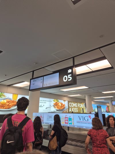 The image depicts a baggage claim area in Schwechat, Austria. A digital display board shows flight information, with "OS 184" to Munich and "OS 772" to Sofia listed. Another sign prominently displays "05" with a luggage icon, indicating carousel number 5. Several individuals are visible, some standing near the baggage carousel and others looking at the displays. Advertisements are present, including one for "VIG Vienna Insurance Group" and another with the slogan "COME FOR A KISS" and images of schnitzel. The environment appears to be an airport terminal, with a suspended ceiling and bright lighting.