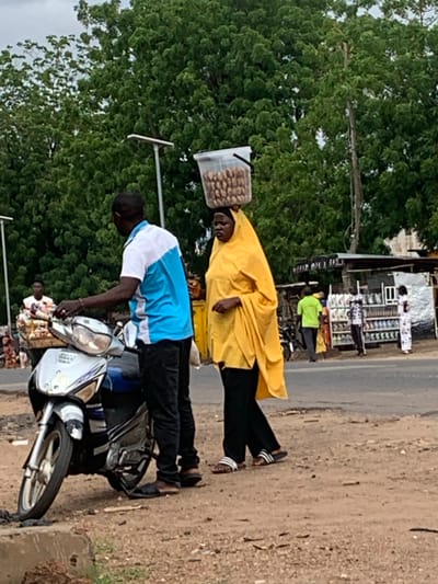 This image captures a daytime street scene in Shendam, Nigeria. In the immediate foreground, a dark-skinned man wearing a white and blue short-sleeved polo shirt, black pants, and brown sandals stands to the left of a silver and blue scooter-style motorcycle. He holds the handlebars of the motorcycle, which is parked on a reddish-brown dirt and gravel shoulder. To his right, a dark-skinned woman, dressed in a bright yellow head covering and matching flowing outer garment over black pants, walks towards the right. She balances a clear plastic bucket filled with numerous light-brown, oval-shaped items (likely nuts or seeds) on her head. Her feet are clad in striped slide sandals.
In the midground, a paved road separates the foreground figures from a row of makeshift market stalls on the opposite side. These stalls are constructed from corrugated metal and wood, displaying various products. Several individuals are visible near these stalls, including a person in a bright green shirt, another in a patterned shirt, and a third in a white and patterned dress. Further left in the midground, another dark-skinned man in a white shirt is partially visible, attending to a wooden cart filled with items. Two modern streetlights, each with a flat, square solar panel, stand on tall poles on the left and center-right portions of the frame. The background consists of dense green foliage from numerous trees, indicating a lush environment under an overcast sky.