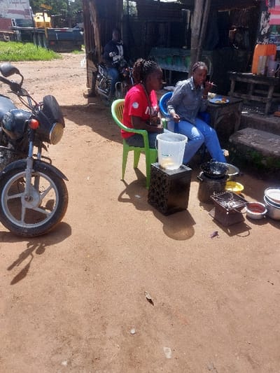 This image depicts an outdoor scene in Kimamba, Tanzania. In the foreground, the front wheel and engine of a dark motorcycle are visible on the left. The ground is packed dirt, marked by distinct shadows indicating sunny conditions.
Two women are seated in the midground. The woman on the left wears a red t-shirt with a white logo and sits on a light green plastic chair. Her hair is styled in braids. The woman on the right wears a grey denim-style button-up shirt and blue jeans, seated on a blue plastic chair. She holds an object to her mouth, appearing to be eating from a small plate on a rustic wooden table behind her. Between the two women, a clear plastic bucket containing light-colored items rests on a black plastic crate. To the right of the bucket, a small charcoal stove with a frying pan on top is present, alongside several metal pots and bowls on the ground.
In the background, a man is seated on a dark motorcycle, positioned near a wooden support post of a basic, shaded structure, likely an outdoor eatery or stall. Further in the distant background on the left, part of a yellow heavy vehicle, possibly an excavator or truck, is visible beyond a patch of green vegetation. The overall structure behind the people consists of wooden posts and a dark, possibly corrugated metal or thatch, roof providing shade.