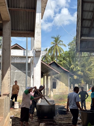 In Kayeepanyang, Indonesia, a group of men are gathered around a large cooking vessel. The men are stirring the contents of the pot with long wooden paddles. Smoke rises from the vessel. Nearby, there is a building with a corrugated metal roof, and in the background, a palm tree and another building can be seen under a partly cloudy sky. A partially constructed building, with metal posts, is in the foreground.