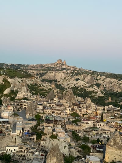 This wide panoramic image presents a captivating view of Göreme, Cappadocia, Turkey, during dusk or early evening. The scene showcases the region's distinctive landscape, characterized by numerous eroded volcanic rock formations, often referred to as "fairy chimneys," which dominate the terrain.

The foreground and midground are densely populated with a town, where buildings are intricately built into and around these natural rock structures. Many dwellings appear to be cave homes, carved directly into the soft stone, while others are constructed from light-colored local materials that blend harmoniously with the surroundings. These structures feature multiple stories, small windows, and flat roofs, reflecting traditional architectural styles. As twilight settles, numerous artificial lights illuminate the buildings, casting a warm glow and indicating active habitation or commercial activity within the town.

Beyond the immediate settlement, the landscape continues with undulating hills and valleys, still dotted with the unique rock formations and occasional smaller clusters of buildings, extending towards the horizon. Sparse green vegetation, including trees and shrubs, is visible, primarily nestled in the valleys and around some of the dwellings, providing a natural contrast to the pale rock.

The sky transitions from a clear, light blue overhead to a softer, pale orange-yellow tint near the horizon, typical of a sunset or twilight period. The absence of clouds suggests clear weather conditions. The image captures a serene and timeless quality, emphasizing the unique blend of natural beauty and historical human ingenuity without depicting any explicit human activity or individuals.