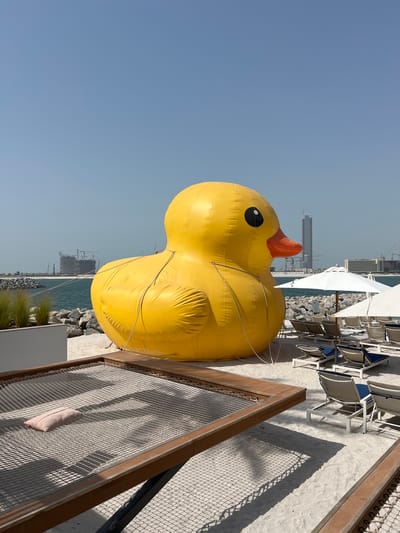 An oversized yellow inflatable duck is the central subject, positioned on a sandy beach. The duck is detailed with a black eye and an orange beak. It is tethered by ropes. In the background, the scene transitions to a body of water, beyond which a cityscape with buildings under construction and cranes is visible. The sky is clear and blue.

The setting appears to be a beach resort, with lounge chairs and umbrellas arranged on the sand. A wooden-framed structure with a mesh surface, possibly part of a seating area or playground, is in the foreground. A light-colored cushion rests on this mesh. The lighting suggests it is daytime, with sunlight casting shadows on the sand and mesh.

The image captures a moment of leisure at a beach, with the large inflatable duck adding a playful and unusual element to the scenery. The presence of construction in the distance hints at ongoing development in the area, possibly indicating a location in a rapidly developing region like the United Arab Emirates. No text is visible in the image.