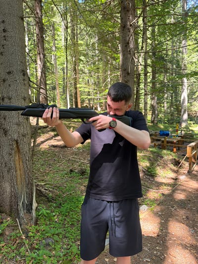 A man in a black t-shirt and black shorts is holding a black rifle in a forest. He is looking through the sights of the rifle. The forest has tall trees and green foliage. In the background, there is a wooden picnic table and a wooden fence. The ground is covered in dirt and fallen leaves. The sun is shining through the trees, creating dappled light and shadow. The scene appears to be in the city of Govedartsi, Bulgaria.