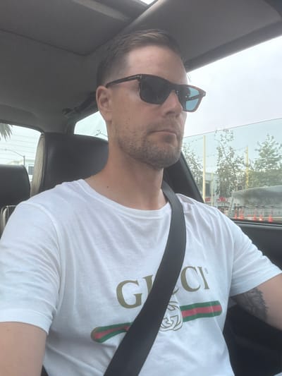 A man with short brown hair and a beard is in the driver's seat of a car. He is wearing sunglasses and a white t-shirt with the Gucci logo. The car appears to be moving, and the background shows trees and buildings in an urban environment. The photo was taken on July 28, 2023, in Santa Monica, California.