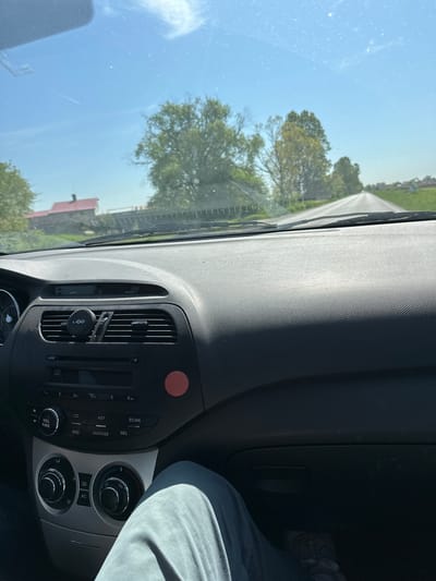 The image captures a driver's perspective from inside a car on a bright, sunny day, likely in Voysil, Bulgaria. The view is primarily of the dashboard and the road ahead through the windshield, which appears somewhat dusty or speckled.

In the foreground and to the right, a significant portion of the dark grey dashboard is visible, textured with a small, repetitive pattern. The central console features a car radio/media unit with several buttons and two rotary knobs for climate control. A red, circular hazard light button is positioned above the radio unit. A phone mount, bearing the "UGO" logo, is clipped into one of the air vents. Partially visible on the far right is a person's lap and leg, dressed in light grey pants, suggesting a passenger or the driver themselves.

Looking out through the windshield, a clear blue sky dominates the upper portion of the exterior view. Scattered trees with green leaves line what appears to be a rural or country road, receding into the distance. A faint structure with a reddish roof can be seen amidst the trees in the mid-ground. The weather is clear and bright, indicating daytime.

Visible text on the car's media console includes: "MUTE", "VOL PWR", "CD", "BND", "AUX/USB", "AST", "SCAN", "SEL", and numbers "2", "3 RPT", "5", "6 DA". The "UGO" brand name is visible on the phone mount. Climate control features include "AC" and fan symbols.