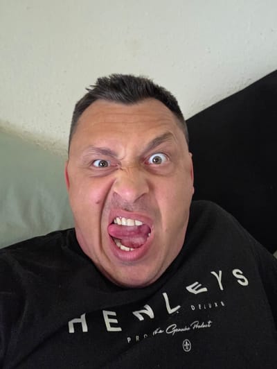 In Shotton, United Kingdom, a close-up, frontal portrait displays an adult male with short, dark hair making a contorted facial expression. His eyes are wide open, and his eyebrows are heavily furrowed, creating prominent wrinkles across his forehead and the bridge of his nose. His mouth is open wide, revealing visible teeth and an extended, downward-curled tongue. He is dressed in a black short-sleeved garment, likely a t-shirt or sweatshirt, which features white printed text. The text prominently displays "HENLEYS DELUXE" in a larger font, with "PRO The Genuine Product" in a smaller cursive script below it, accompanied by a fleur-de-lis symbol. The background consists of a light-colored, textured surface on the left side of the frame and a dark, indistinct area on the right, separated by a diagonal line. The lighting is even, suggesting an indoor environment.