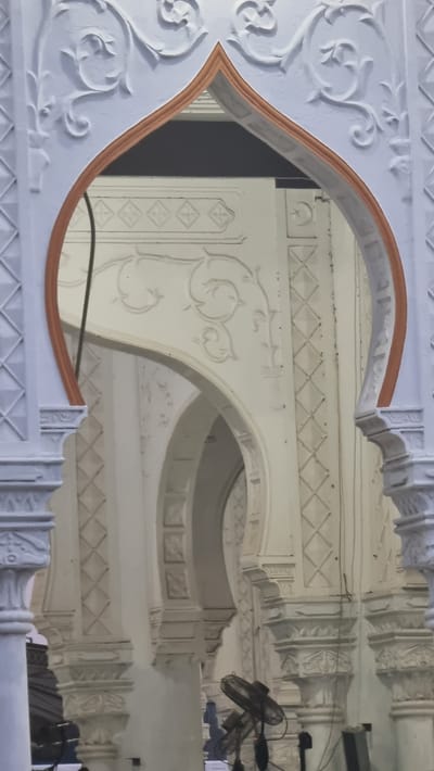 The image shows a close-up view of an architectural detail, likely part of a mosque or a similar traditional building, in Kambam, Indonesia. The foreground features a white, intricately carved archway with decorative scrollwork and geometric patterns. The arch is framed by a thin, reddish-brown trim. Beyond this arch, the scene opens up to reveal more of the building's interior, characterized by multiple cream-colored arches and columns. These columns are adorned with relief carvings, including diamond shapes and floral motifs. The lighting is subdued, creating a sense of depth and highlighting the textures of the stone or plasterwork. In the background, two electric fans are visible, suggesting the presence of people and the need for ventilation in the space. The overall impression is one of ornate, traditional Islamic architecture, with a touch of practicality in the form of the fans.