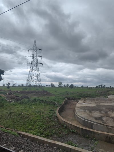 The image depicts a cloudy, overcast day with a prominent electricity pylon dominating the center-left of the frame. The pylon is tall and metallic, with numerous wires extending from it. The surrounding landscape consists of green fields with scattered trees and some rocky, uneven terrain in the foreground. A concrete structure with curved edges is visible on the right side of the image. Train tracks are present at the bottom of the frame. The location is the city of Khandwa, India.