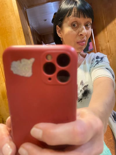 A woman with dark hair and bangs is taking a selfie with a red smartphone. She is sticking her tongue out playfully. She is wearing a white sweater with a black floral pattern. The background appears to be a hallway with wooden walls. The image does not contain any visual information that allows for the determination of its location within the city of Yokohama, Japan.