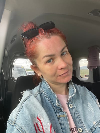 A woman with red hair is sitting in the passenger seat of a car. She is wearing a light pink t-shirt and a light blue denim jacket with red and white embroidery on the sleeve. Her sunglasses are on top of her head. The car interior is visible, including the dark seats and the car's ceiling. Through the car window, a slightly overcast sky and some distant greenery can be seen. The image appears to be taken indoors, possibly in a parking lot or on a road. The woman's expression is neutral.