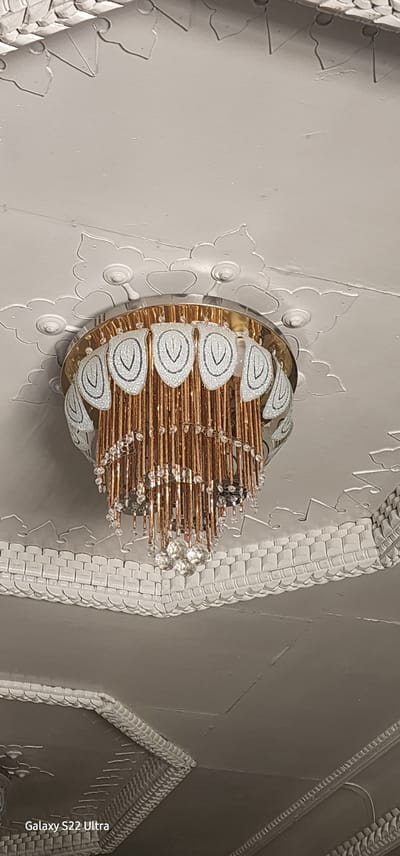 An ornate chandelier hangs from a white, textured ceiling adorned with decorative plaster molding. The chandelier features a gold-toned rim, a band of glass panels decorated with white and black paisley-like designs, and numerous dangling strands of small, copper-colored rods and beads. A faceted crystal ornament hangs from the center of the chandelier. The ceiling molding is intricate, with floral and basket-weave patterns. The scene appears to be indoors, likely in a residential or public building, in Tashkent, Uzbekistan.