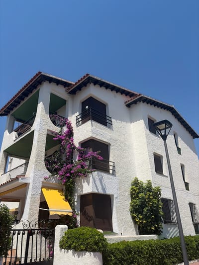 The image shows a white stucco building with a red tile roof under a clear blue sky. The building has several balconies with black wrought iron railings and dark shutters on the windows. Purple bougainvillea is climbing up the side of the building, trailing over balconies. A yellow awning is extended over a ground-floor window or doorway on the left side of the building. In front of the building, there is a black wrought iron fence with decorative scrollwork, and a neatly trimmed green hedge runs along the base of the fence and the building. A dark grey lamppost stands to the right of the building. The environment appears to be a sunny day in a coastal town. While the specific street or exact location within Roda de Berà, Spain is not identifiable from the image alone, the architectural style and vegetation are consistent with a Mediterranean setting. There are no people or animals visible in the image.