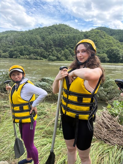 Two individuals are standing by a river, ready for rafting. The person on the left, wearing glasses, a white long-sleeved shirt, a yellow life jacket, and purple leggings, holds a paddle. The person on the right, with curly brown hair and a yellow helmet, wears a yellow life jacket and black shorts, also holding a paddle. They are positioned near lush green vegetation and a calm river, with a densely forested hillside in the background under a cloudy sky. The overall scene suggests an outdoor recreational activity.