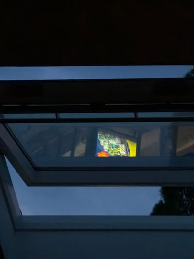 The image shows a reflection of a colorful advertisement on a skylight. The advertisement appears to be for a store or event, with text and images that are not entirely legible. The skylight is open and angled, allowing a view of the sky and some trees in the background. The overall scene is dark, suggesting it is either nighttime or early morning. The advertisement's reflection is the most prominent and detailed element in the image. There is no indication of the specific location in Witten, Germany, nor any people or actions depicted.